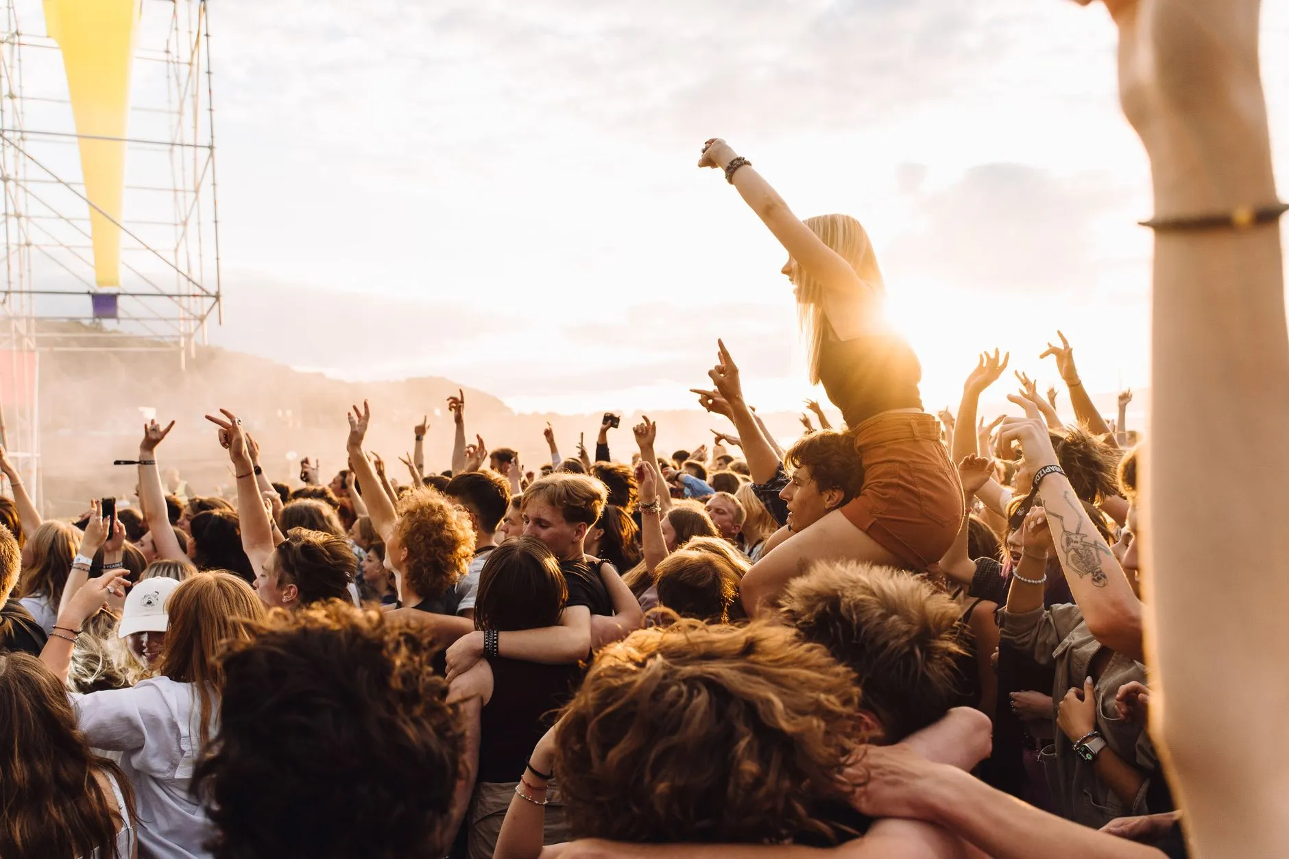 Plateia de festival ao ar livre com os braços erguidos contra o sol do fim de tarde, uma moça nos ombros de um amigo